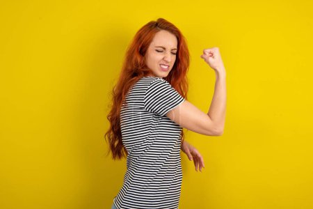 Photo for Portrait of funny red haired woman wearing striped shirt over yellow studio background shout yeah raise fists hands celebrate victory game competition - Royalty Free Image