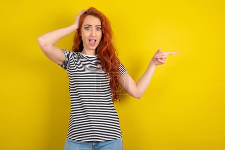 Photo for Young beautiful red haired woman wearing striped shirt over yellow studio background pointing at empty space holding hand on head - Royalty Free Image