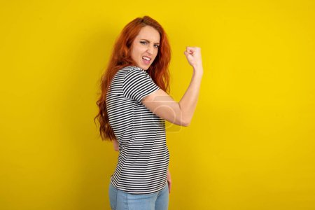Téléchargez les photos : Jeune belle femme aux cheveux rouges portant une chemise rayée sur fond jaune studio célèbre la victoire - en image libre de droit