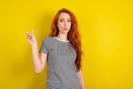 Téléchargez les photos : Femme aux cheveux rouges portant une chemise rayée sur fond jaune studio fait geste de paix garde les lèvres pliées montre v signe. Concept de langage corporel - en image libre de droit