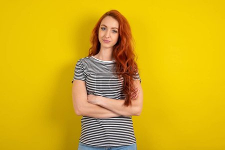 Téléchargez les photos : Femme aux cheveux rouges confiante portant une chemise rayée sur fond de studio jaune avec les bras croisés regardant vers la caméra - en image libre de droit