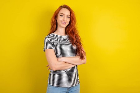 Téléchargez les photos : Portrait de femme aux cheveux rouges portant une chemise rayée sur fond jaune studio debout avec les bras croisés et souriant - en image libre de droit