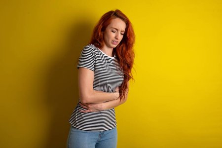 Photo for Red haired woman wearing striped shirt over yellow studio background got stomachache - Royalty Free Image