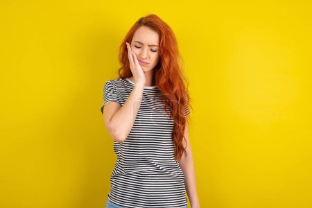 Photo for Red haired woman wearing striped shirt over yellow studio background with toothache on yellow background - Royalty Free Image
