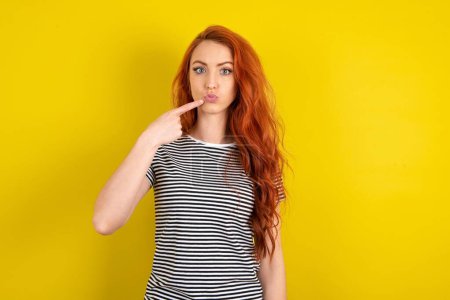 Foto de Encantadora mujer de pelo rojo con camisa a rayas sobre fondo amarillo estudio apuntando en los labios de la puchera con el dedo índice, mostrando efecto después del procedimiento de elevación - Imagen libre de derechos