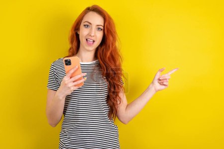 Téléchargez les photos : Femme aux cheveux rouges étonnée portant une chemise rayée sur fond jaune studio tenant son téléphone et pointant du doigt à côté de l'espace de copie vide - en image libre de droit