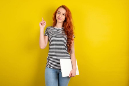 Téléchargez les photos : Femme aux cheveux rouges portant une chemise rayée sur fond de studio jaune tenant un ordinateur portable et montrant sur un espace vide - en image libre de droit