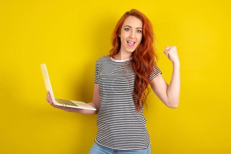 Téléchargez les photos : Femme aux cheveux rouges portant une chemise rayée sur fond jaune studio tenir ordinateur bouche ouverte lever poing - en image libre de droit