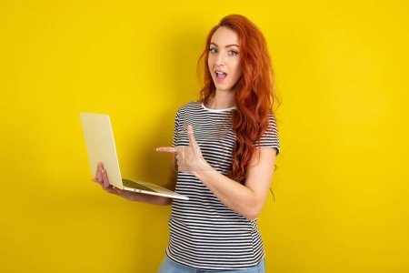 Photo for Shocked red haired woman wearing striped shirt over yellow studio background pointing finger modern device - Royalty Free Image