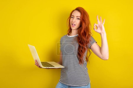 Photo for Attractive cheerful skilled red haired woman wearing striped shirt over yellow studio background using laptop showing ok-sign winking - Royalty Free Image