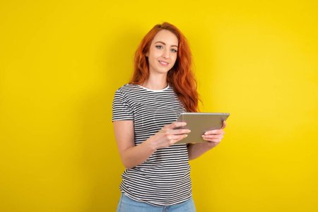 Foto de Foto de mujer optimista de pelo rojo con camisa a rayas sobre la tableta de retención de fondo de estudio amarillo - Imagen libre de derechos