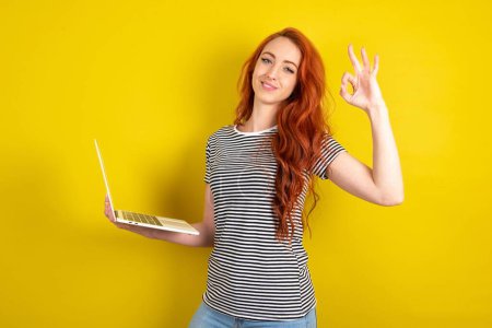 Photo for Positive red haired woman wearing striped shirt over yellow studio background hold wireless netbook with hand fingers show okey symbol - Royalty Free Image