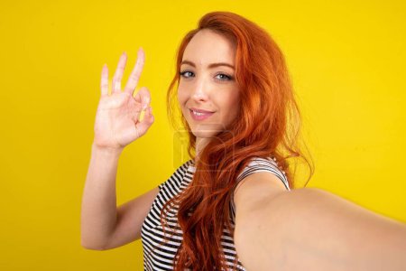 Photo for Portrait of pretty cheerful red haired woman wearing striped shirt over yellow studio background make selfie show okey symbol - Royalty Free Image