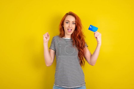 Photo for Photo of lucky impressed red haired woman wearing striped shirt over yellow studio background with arm fist holding credit card. Celebrated - Royalty Free Image