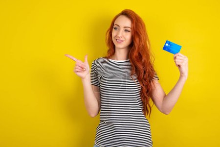 Photo for Curious smiling red haired woman wearing striped shirt over yellow studio background showing plastic bank showing finger copyspace - Royalty Free Image