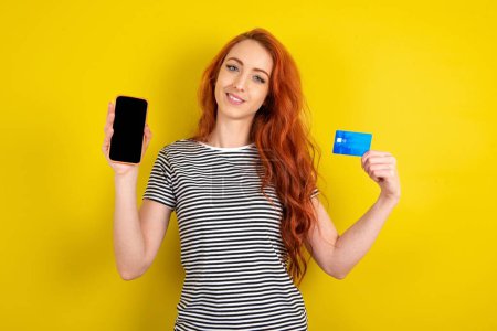 Photo for Red haired woman wearing striped shirt over yellow studio background opened bank account, holding smartphone and credit card, smiling, recommend use online shopping application - Royalty Free Image