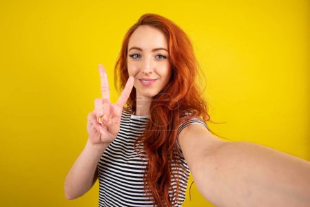 Téléchargez les photos : Positif cheveux rouges femme aux cheveux rouges portant chemise rayée sur fond jaune studio portant chemise rayée sur fond jaune studio prendre selfie faire v-signe - en image libre de droit