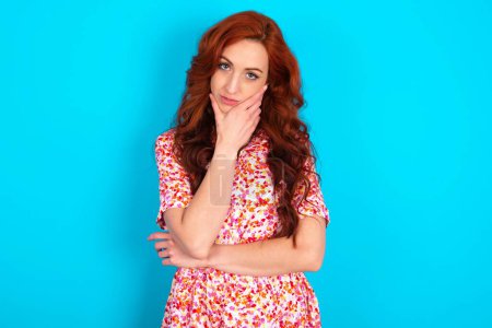 Photo for Thoughtful smiling Young redhead woman wearing floral dress over blue background keeps hand under chin, looks directly at camera, listens something with interest. Youth concept. - Royalty Free Image