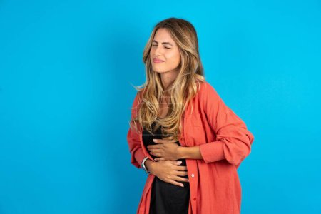 blonde woman wearing overshirt  with hand on stomach because nausea, painful disease feeling unwell. Ache concept.