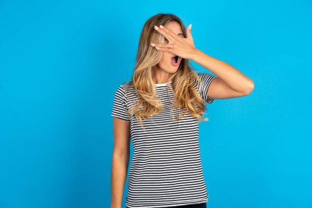 Photo for Young beautiful woman wearing striped t-shirt over blue background peeking in shock covering face and eyes with hand, looking through fingers with embarrassed expression. - Royalty Free Image