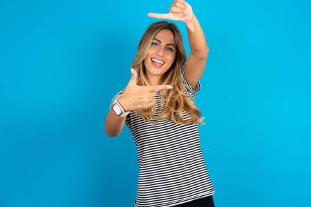 Photo for Young beautiful woman wearing striped t-shirt over blue background making finger frame with hands. Creativity and photography concept. - Royalty Free Image