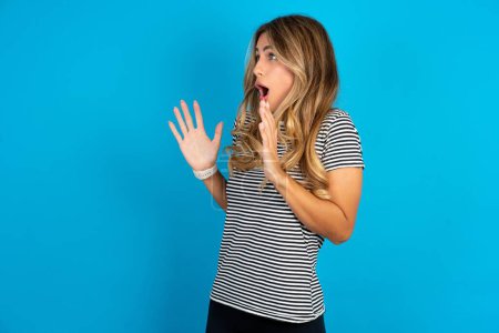 Photo for Young beautiful woman wearing striped t-shirt over blue background shouts loud, keeps eyes opened and hands tense. - Royalty Free Image