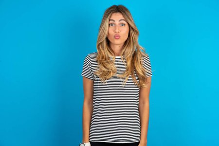 Photo for Shot of pleasant looking young beautiful woman wearing striped t-shirt over blue background, pouts lips, looks at camera, Human facial expressions - Royalty Free Image