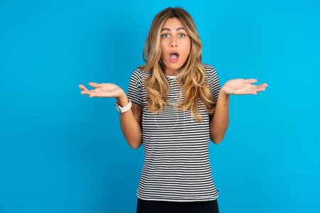 Photo for Frustrated young beautiful woman wearing striped t-shirt over blue background feels puzzled and hesitant, shrugs shoulders in bewilderment, keeps mouth widely opened, doesn't know what to do. - Royalty Free Image