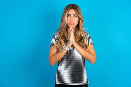 Photo for Positive young beautiful woman wearing striped t-shirt over blue background smiles happily, glad to receive pleasant news from interlocutor, keeps palms together, People emotions concept. - Royalty Free Image
