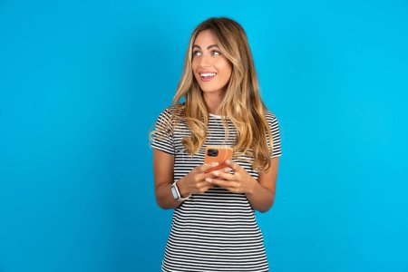 Foto de Joven hermosa mujer vistiendo camiseta a rayas sobre fondo azul sosteniendo un teléfono inteligente y mirando hacia los lados en blanco copyspace. - Imagen libre de derechos