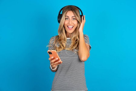 Photo for Positive young beautiful woman wearing striped t-shirt over blue background  holds modern cell phone connected to headphones, clenches fist from good emotions, exclaims with joy, - Royalty Free Image