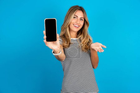 Photo for Young beautiful woman wearing striped t-shirt over blue background with a mobile. presenting smartphone. Advertisement concept. - Royalty Free Image