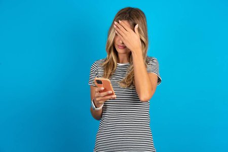 Photo for Young beautiful woman wearing striped t-shirt over blue background looking at smart phone feeling sad holding hand on face. - Royalty Free Image