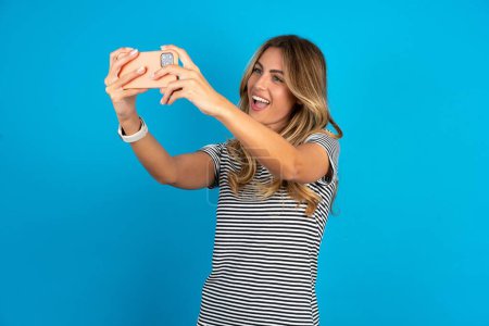 Téléchargez les photos : Jeune belle femme portant un t-shirt rayé sur fond bleu prendre un selfie pour le poster sur les médias sociaux ou avoir un appel vidéo avec des amis. - en image libre de droit