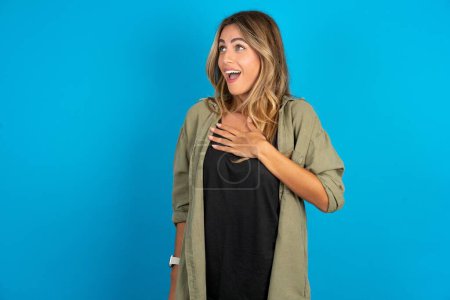 Photo for Joyful beautiful blonde woman wearing overshirt on blue background expresses positive emotions recalls something funny keeps hand on chest and giggles happily. - Royalty Free Image