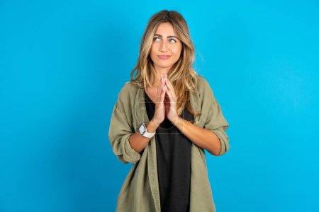 Photo for Beautiful blonde woman wearing overshirt on blue background steepled fingers and looks mysterious aside has great evil plan in mind - Royalty Free Image