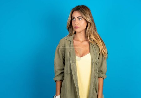 Photo for Beautiful blonde woman wearing overshirt on blue background stares aside with wondered expression has speechless expression. Embarrassed girl looks in surprise - Royalty Free Image
