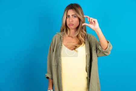 Photo for Beautiful blonde woman wearing overshirt on blue background purses lip and gestures with hand, shows something very little. - Royalty Free Image