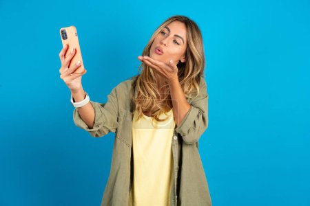 Téléchargez les photos : Belle femme blonde portant overshirt sur fond bleu souffle baiser d'air à la caméra du smartphone et prend selfie, envoie mwah via appel en ligne. - en image libre de droit