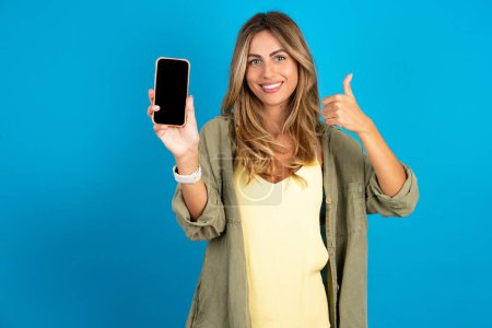 Foto de Hermosa mujer rubia vistiendo overshirt sobre fondo azul Mostrar pantalla en blanco smartphone, pulgar hacia arriba recomendar nueva aplicación - Imagen libre de derechos