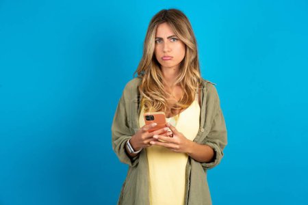 Photo for Upset dissatisfied beautiful blonde woman wearing overshirt on blue background uses mobile software application and surfs information in internet, holds modern mobile hand - Royalty Free Image