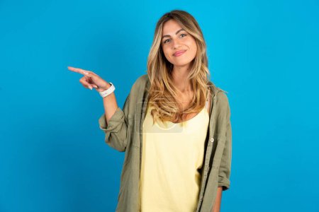beautiful blonde woman wearing overshirt on blue background points to side on blank space demonstrates advertisement. People and promotion concept