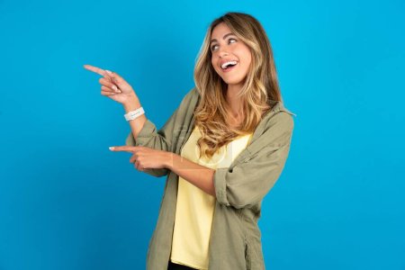 Optimistic beautiful blonde woman wearing overshirt on blue background points with both hands and  looking at empty space.