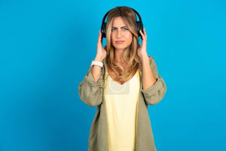 Photo for Serious displeased beautiful blonde woman wearing overshirt on blue background looks puzzled at camera being angry wears stereo headphones listens music while walking at street - Royalty Free Image