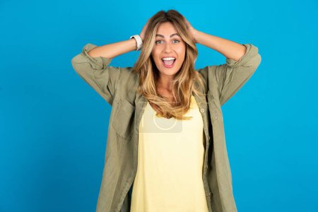 Photo for Cheerful overjoyed beautiful blonde woman wearing overshirt on blue background reacts rising hands over head after receiving great news. - Royalty Free Image