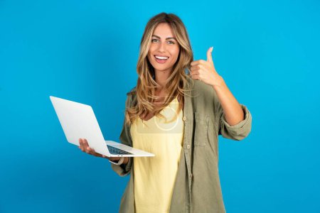Beautiful attractive young beautiful blonde woman wearing overshirt on blue background hold netbook hand finger show thumb up