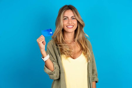 Photo for Photo of happy cheerful smiling positive young beautiful blonde woman wearing overshirt on blue background recommend credit card - Royalty Free Image