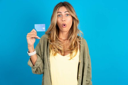 Foto de Foto de una joven hermosa mujer rubia que usa una camiseta sobre fondo azul sorprendida celebrar el pago de la tarjeta de crédito - Imagen libre de derechos