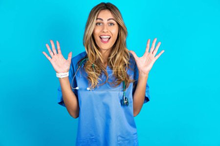 Photo for Optimistic beautiful doctor woman standing over blue studio background raises palms from joy, happy to receive awesome present from someone, shouts loudly, - Royalty Free Image