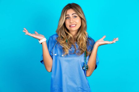 Photo for Clueless beautiful doctor woman standing over blue studio background shrugs shoulders with hesitation, faces doubtful situation, spreads palms, Hard decision - Royalty Free Image